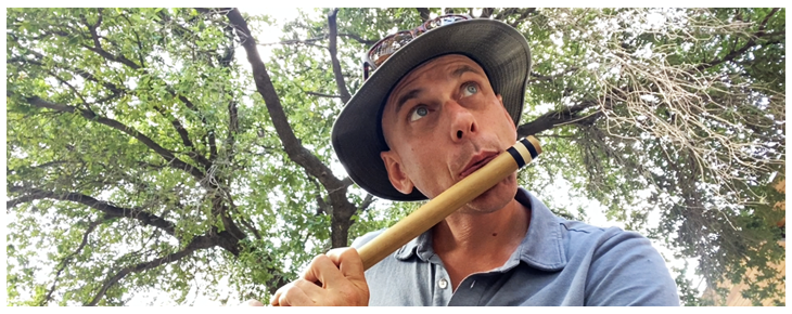 John Davison playing a wooden flute under a tree